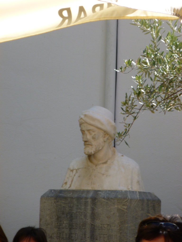 White stone bust on a grey-green stone plinth.  Al-Ghafiqi is also wearing a cloth cap and a robe.
